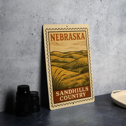 a metal sign with a vintage-style illustration of a rural landscape, including rolling hills and a field, with the text "NEBRASKA SANDHILLS COUNTRY" prominently displayed.