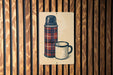 A metal sign with a plaid pattern and a white mug is displayed against a wooden wall.