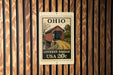 A metal sign with a wooden background, featuring an illustration of a covered bridge and the text "OHIO COVERED BRIDGE USA 20c".