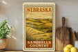 a vintage-style metal sign with the text "NEBRASKA SANDHILLS COUNTRY" displayed prominently. The sign is mounted on a wall, and there are some kitchen utensils, such as a wooden cutting board and a knife, as well as a potted plant and a bowl of fruit, including lemons, in the background.