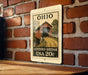 A metal sign with a wooden bridge and the text "OHIO COVERED BRIDGE USA 20c" is displayed on a brick wall.