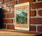 A metal sign with a vintage-style illustration of a mountain road, mounted on a brick wall.