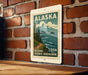 A vintage-style postage stamp depicting Alaska, with a mountain landscape and ocean scene, is displayed on a brick wall.