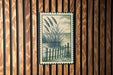 A framed illustration of tall grasses and a wooden fence is displayed on a wooden wall.