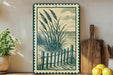 a framed artwork depicting a beach scene with a wooden fence and grassy plants, placed on a shelf next to a cutting board and a potted plant.