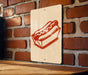 A sign featuring a red hot dog in a cardboard box is mounted on a brick wall.