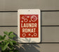 A red and white sign with the text "LUNDROMAT" is mounted on a gray wall, with a plant visible to the left.