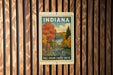 A vintage-style metal sign with the word "INDIANA" and an illustration of a scenic fall landscape with trees and a lake, mounted on a wooden wall.