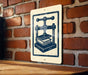 a sign with a black and white illustration of a book press, mounted on a brick wall.