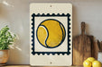 a white sign with a tennis ball design, placed on a wooden surface next to a potted plant and a wooden cutting board.