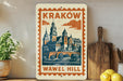 A decorative metal sign with an illustration of a castle or palace, featuring the text "Krakow Wawel Hill" on it, is displayed on a shelf next to a wooden cutting board and some potted plants.