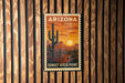 A vintage Arizona Forever USA postage stamp is displayed on a wooden wall, featuring a desert landscape with a cactus and a sunset.