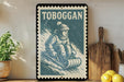 A framed vintage-style stamp with a winter scene depicting a person sledding down a snowy hill, surrounded by kitchen utensils and a potted plant.