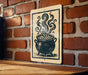 A sign with a black and white illustration of a cauldron with steam rising from it, mounted on a brick wall.