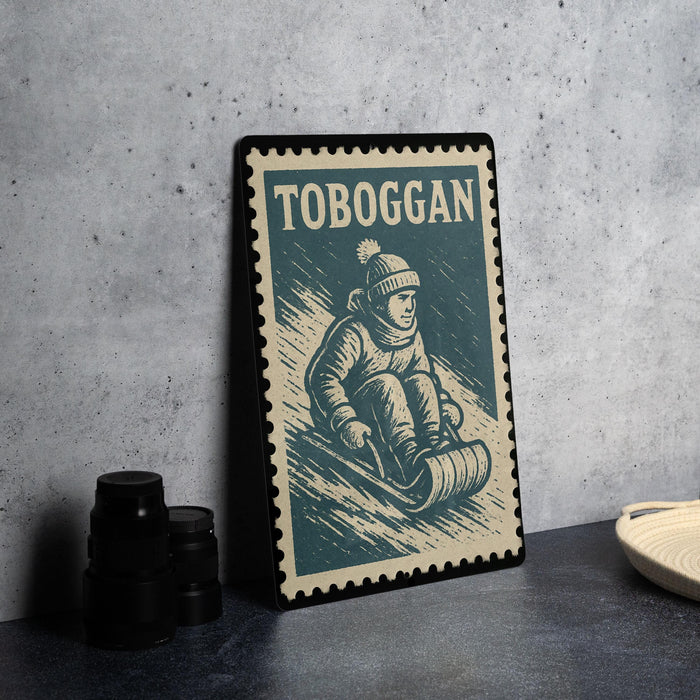 A framed vintage postage stamp depicting a person riding a sled, with the word "TOBOGAN" printed on it.