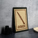 a stamp-like sign with the word "OBOE" printed on it, placed on a table next to some black camera lenses.