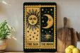 a decorative sign with the sun and moon depicted on it, placed on a shelf next to a wooden cutting board and some potted plants.