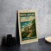 A framed poster of a beach scene with a wooden walkway and ocean waves, sitting on a counter next to some black camera lenses.