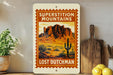 a vintage-style sign for "Superstition Mountains Lost Dutchman", with a desert landscape and a cactus on the sign. The sign is displayed on a shelf next to a wooden cutting board and a potted plant.