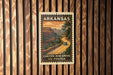 A vintage postage stamp from Arkansas depicting a scenic road leading to a canyon, mounted on a wooden wall.