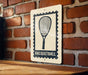 A sign with a racquet on it is mounted on a brick wall.