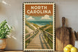 A vintage-style metal sign with the text "NORTH CAROLINA" and an illustration of a wooden boardwalk leading to a beach, surrounded by potted plants and kitchen utensils.
