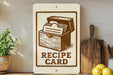 a sign that reads "RECIPE CARD" with a graphic of a recipe book, placed on a shelf next to a wooden cutting board and some kitchen utensils.