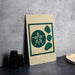 a beige sign with a green design, including a flower and four seashells, placed on a gray countertop next to a stack of black containers.