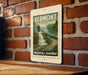 A vintage-style postage stamp depicting a waterfall is mounted on a brick wall, with the words "Vermont" and "Waterfall Parkway" printed below it.