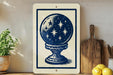 a white sign with a blue illustration of a starry globe, placed on a wooden surface next to a wooden cutting board and a potted plant.