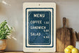 a menu sign with prices for different food items, such as coffee, sandwich, and salad, placed on a wooden countertop next to a potted plant and a cutting board.