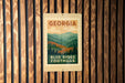A sign for Blue Ridge Foothills in Georgia, featuring a mountain landscape with pine trees and a cloudy sky.