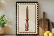 a framed illustration of a knife on a wooden surface, with a potted plant to the left and a wooden cutting board to the right.