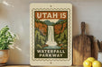 A vintage-style sign for Utah 15 Waterfall Parkway, featuring a waterfall and lush greenery, is displayed on a shelf next to a wooden cutting board and some potted plants.
