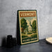 A vintage-style metal sign for Vermont Village Greens, featuring a church and a park scene, is displayed on a gray countertop next to some black camera lenses.