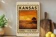 A Kansas sunset stamp is displayed on a shelf, with a wooden cutting board and a potted plant nearby.