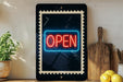 a neon sign that says "OPEN" in red and blue colors, displayed on a black frame. The sign is placed on a wooden surface, with a potted plant, a wooden cutting board, and some lemons nearby.