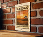 A metal sign with a sunset landscape and the text "KANSAS SUNSET Vista Point" is displayed on a brick wall.