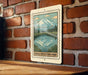 A metal sign with a mountain landscape and a lake is hanging on a brick wall.