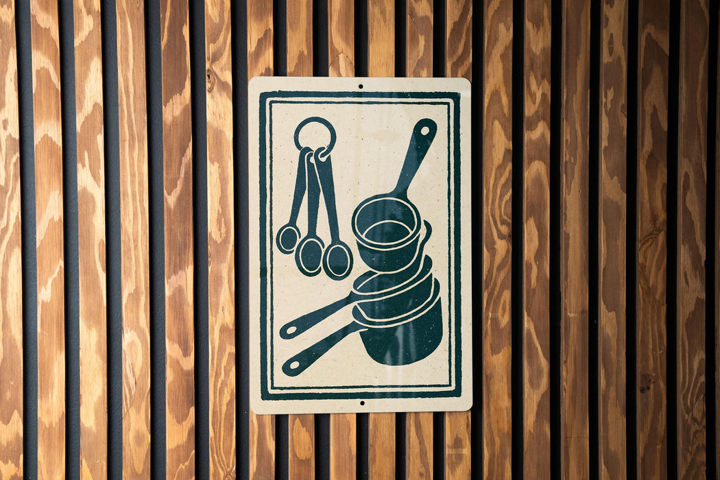 A sign with a pictogram of kitchen utensils, including measuring cups and spoons, mounted on a wooden wall.