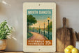 A framed poster of a scenic riverwalk promenade in North Dakota, featuring a path lined with trees and lampposts, with a sunset in the background.