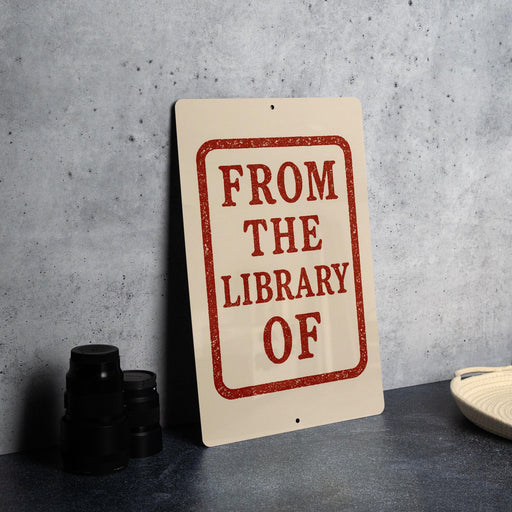 a sign that reads "FROM THE LIBRARY OF" in bold red letters, placed on a gray countertop next to a stack of black camera lenses.