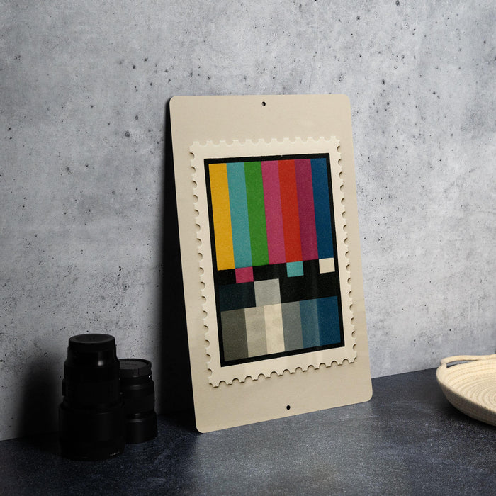 a framed stamp-like artwork with a colorful abstract design, placed on a dark surface next to some black camera lenses.