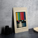 a framed stamp-like artwork with a colorful abstract design, placed on a dark surface next to some black camera lenses.