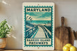 A framed poster of a wooden boardwalk path leading to a beach, with the text "Maryland Barrier Island Pathways" on it, is displayed on a wall next to a wooden cutting board and some potted plants.