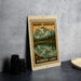 A framed print of a mountain landscape with a lake in the foreground, placed on a countertop next to some black camera lenses.