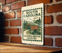 A metal sign with a vintage-style illustration of a covered bridge in the South Carolina countryside, mounted on a brick wall.
