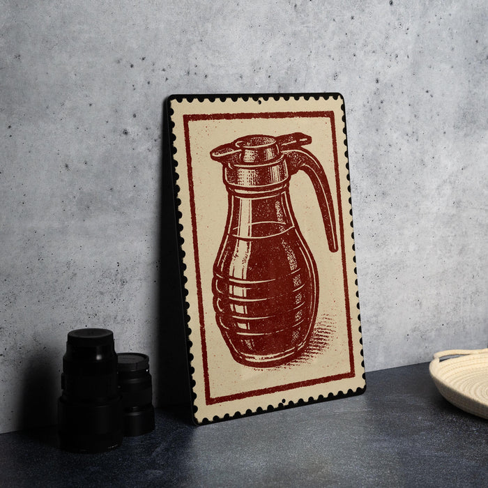 A framed illustration of a coffee pot is displayed on a countertop, accompanied by a stack of black containers and a woven basket.