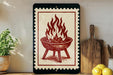 a framed illustration of a grill with flames on it, placed on a shelf next to a wooden cutting board and some lemons.