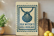 a decorative sign with a seashell and the text "SEA WITCH'S SHELL NECKLACE" on a shelf, accompanied by a potted plant, a wooden cutting board, and some lemons.